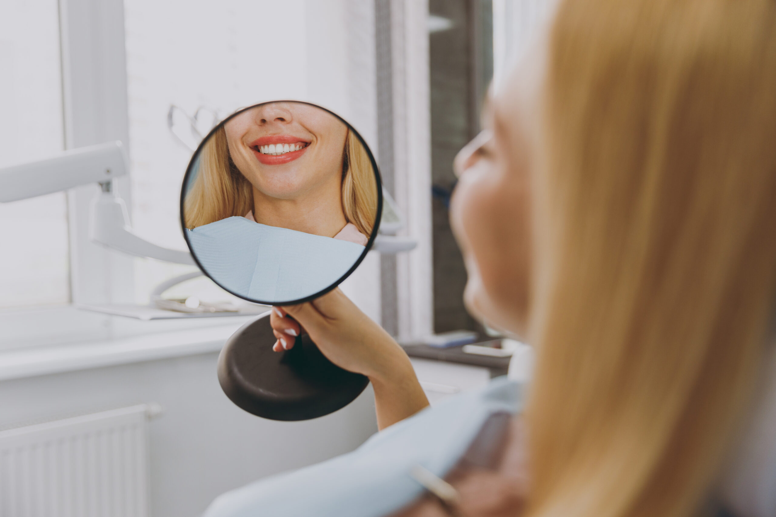 Young smiling satisfied happy woman 20s reflected in mirror sitting at dental office chair indoor light cabinet looking at teeth toothy smile whitening result blink Healthcare caries enamel treatment.