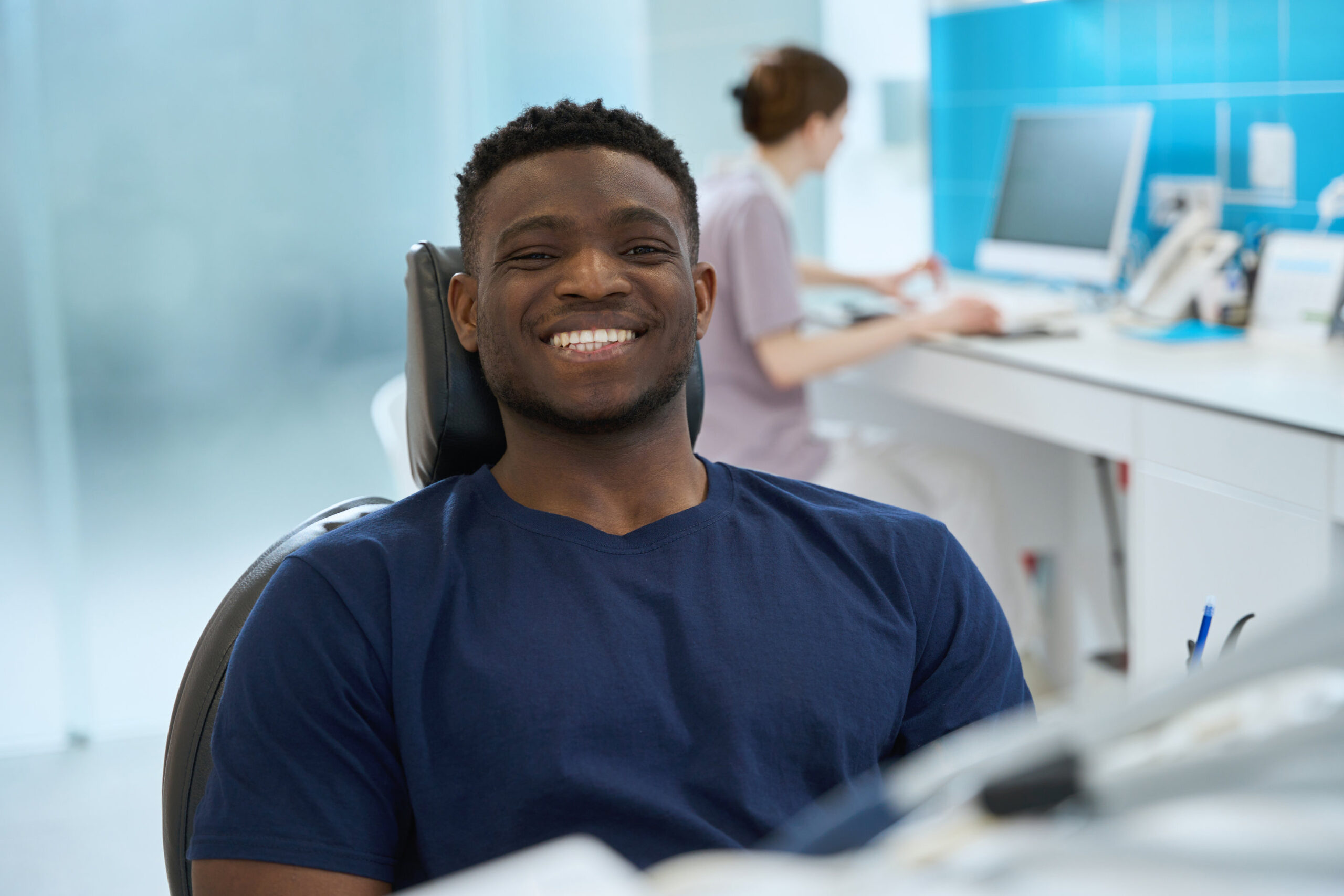 Male client sitting in dental chair with healthy and clean smile while stomatologist keeping medical records on computer