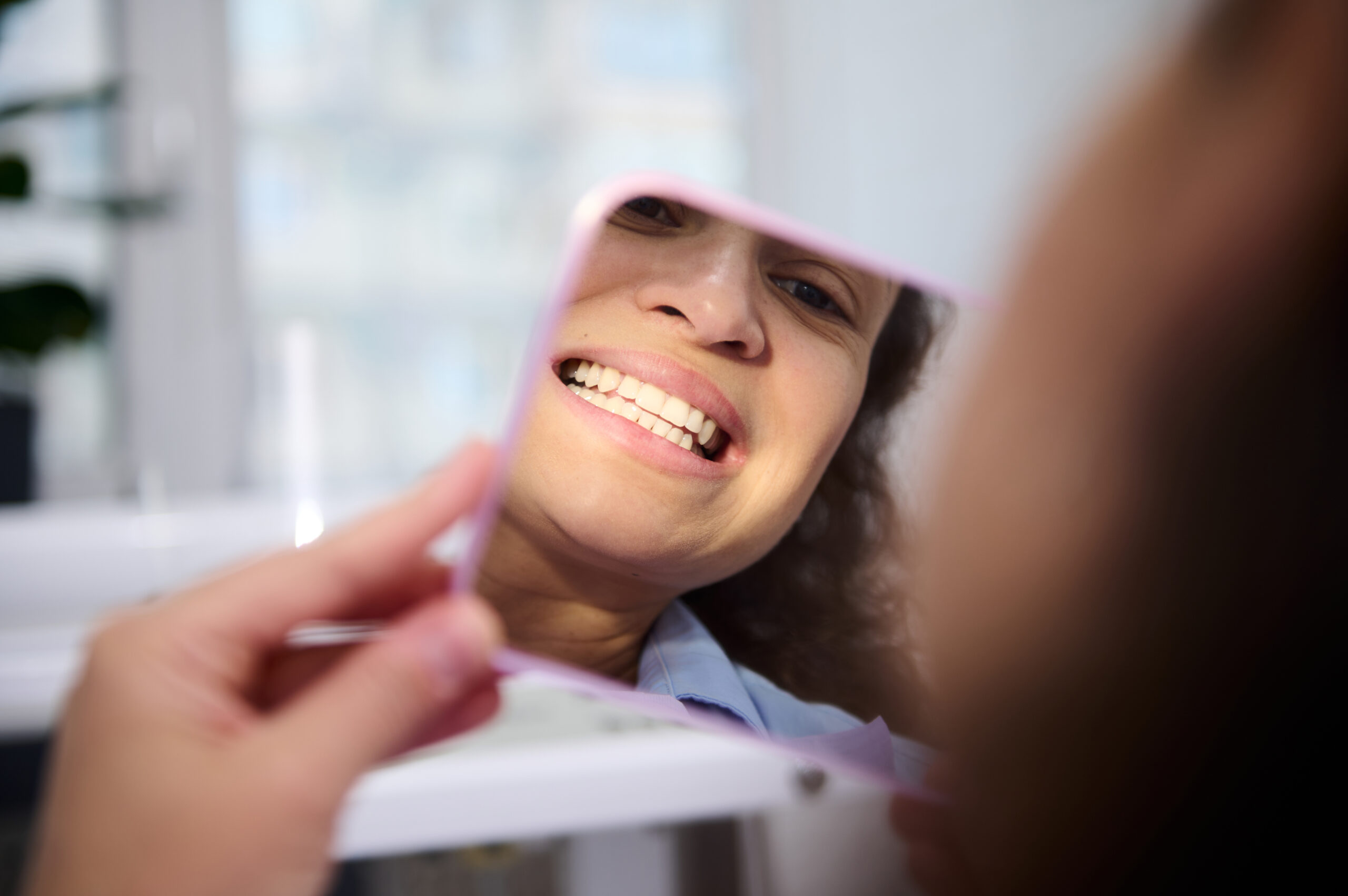 Close-up reflection in the cosmetic mirror of a pretty woman, female patient sitting in dental chair, admiring her beautiful smile and teeth after teeth bleaching procedure in dentistry clinic.
